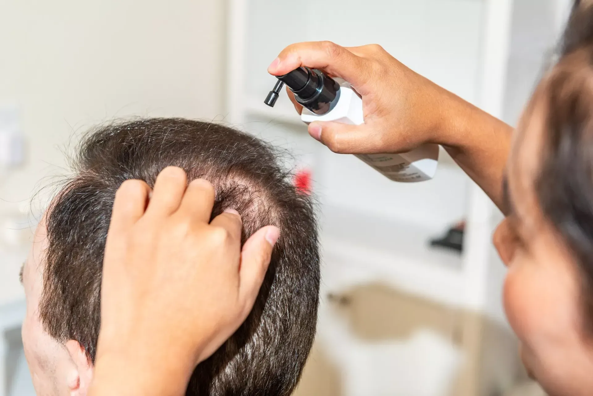 A woman is spraying lotion on a man 's head.