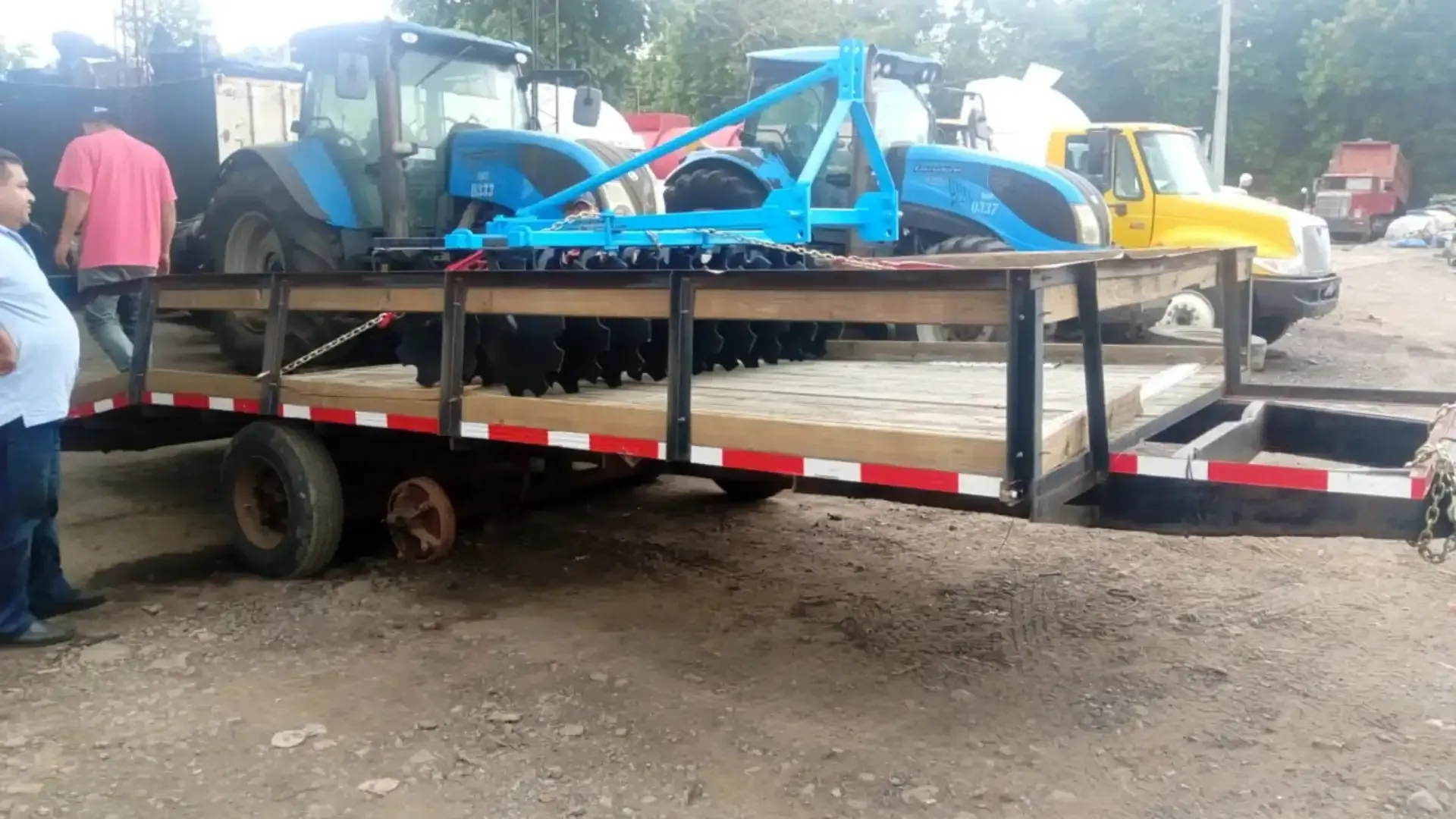 Un hombre está parado junto a un remolque con un tractor azul.