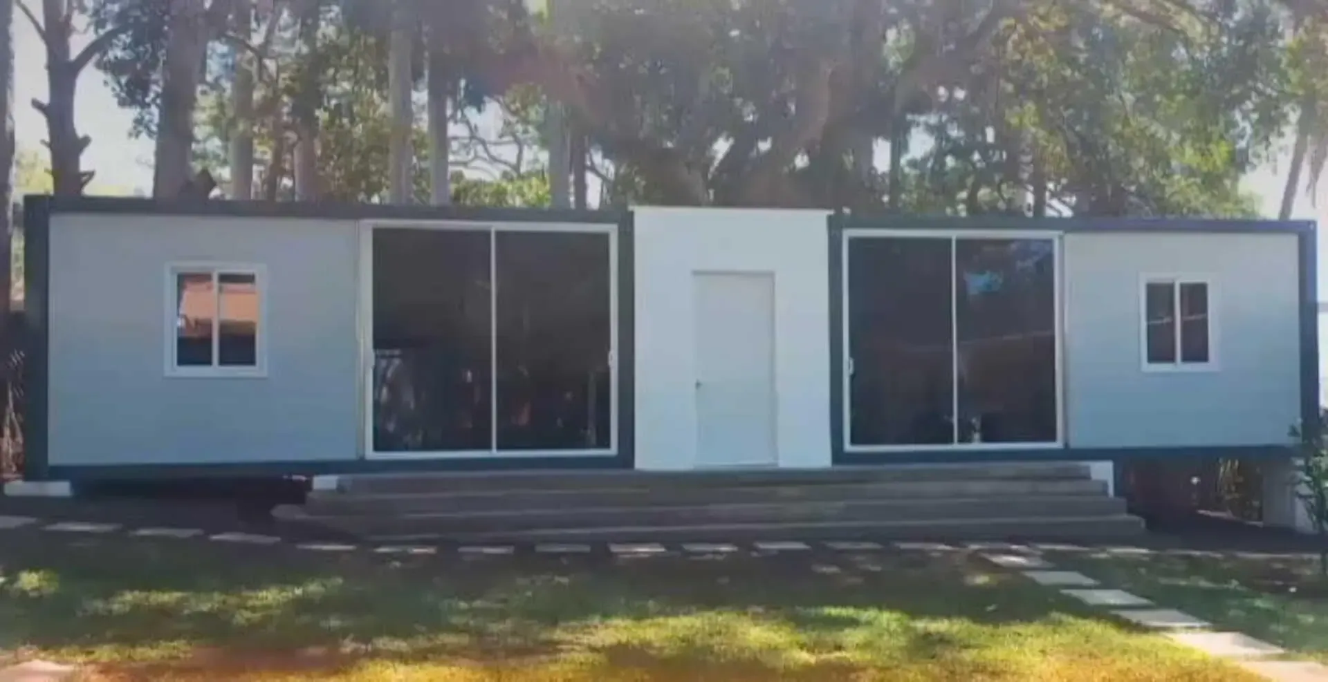 Una casa gris y blanca con puertas corredizas de vidrio se encuentra en lo alto de un exuberante campo verde.