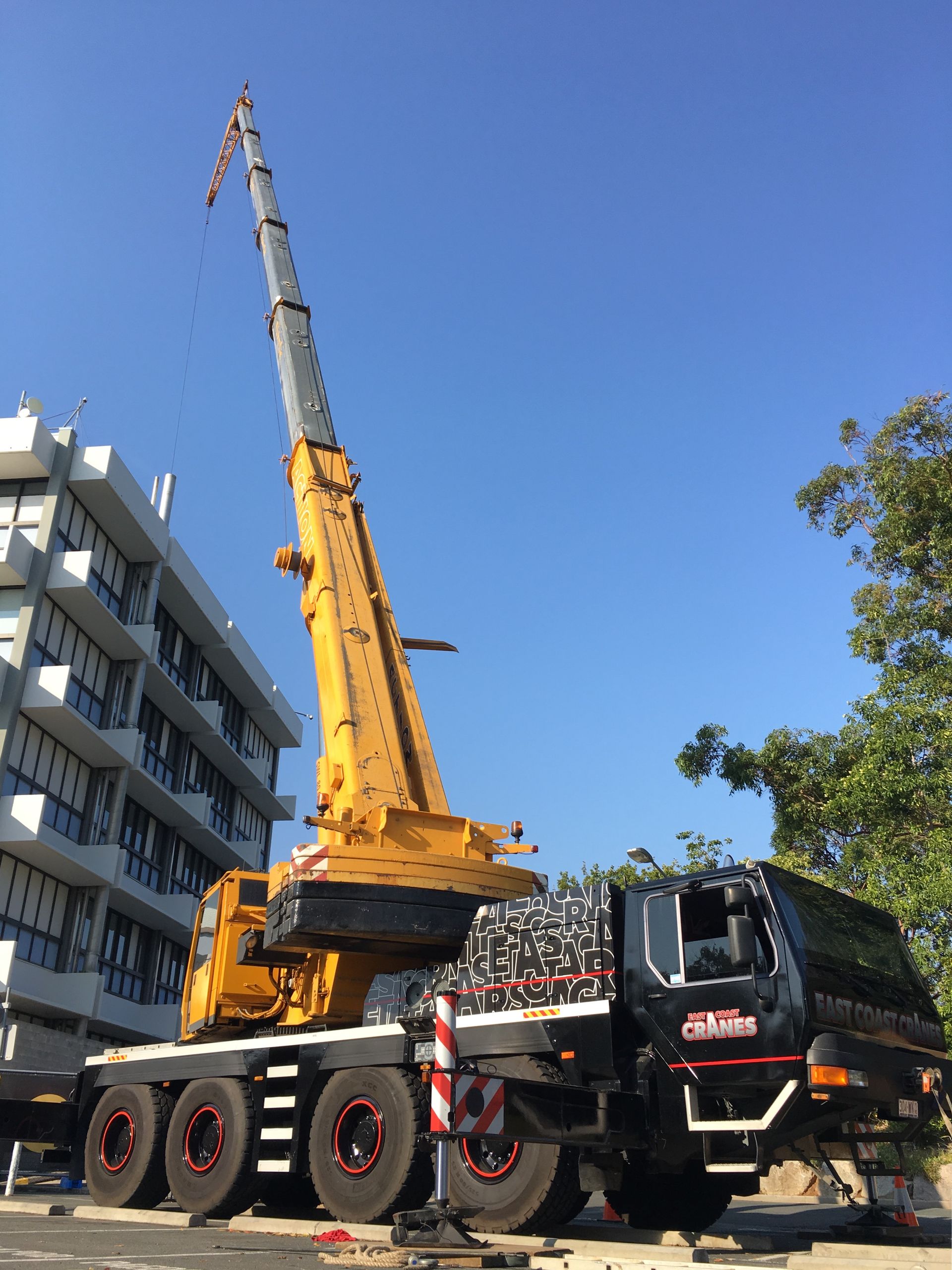 A Large Yellow Crane, Black Cab, Raised Against a Blue Sky, Beside a Building — East Coast Cranes in Ormeau, QLD