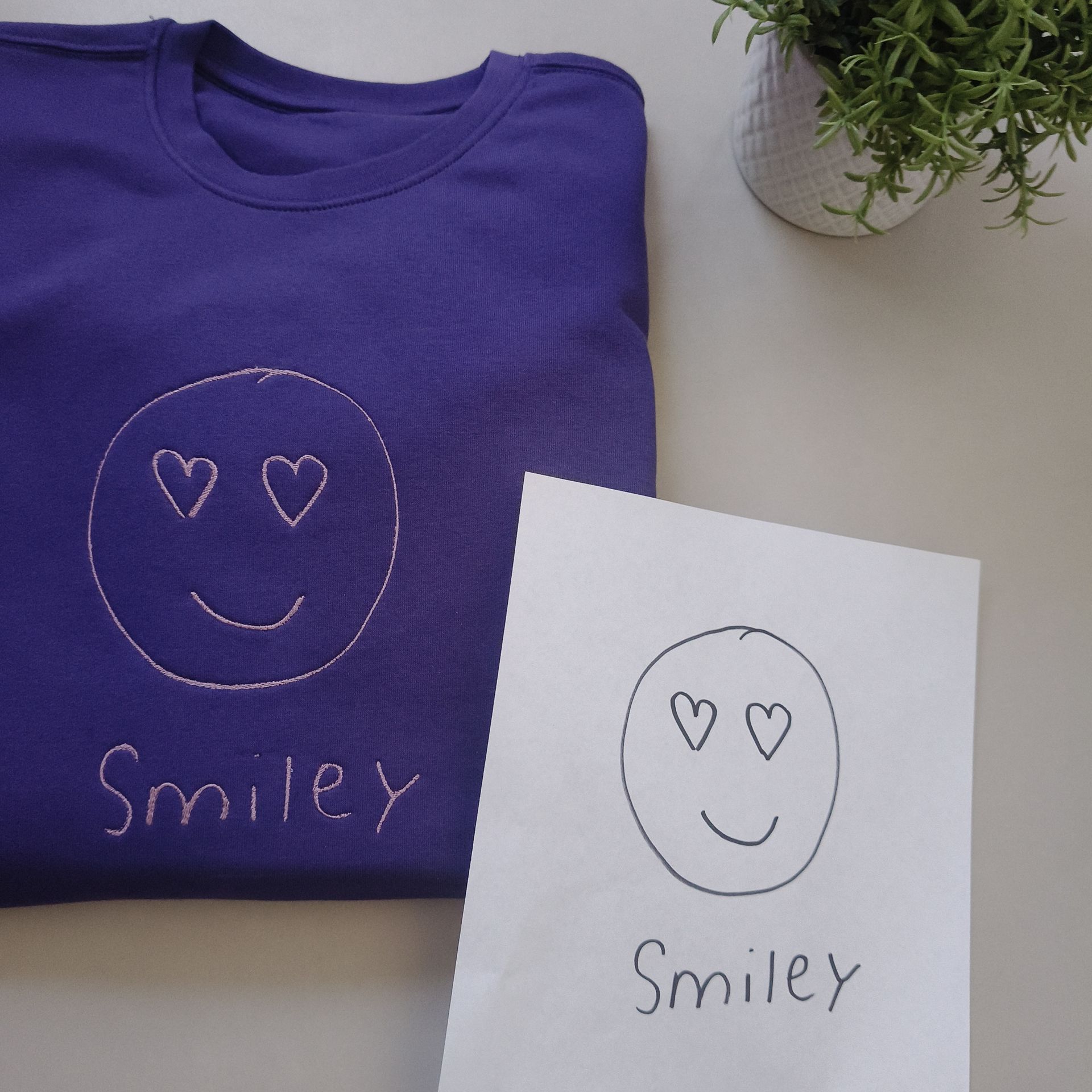 Paarse trui met geborduurd smileygezichtje met hartjesoogjes, naast de originele tekening.