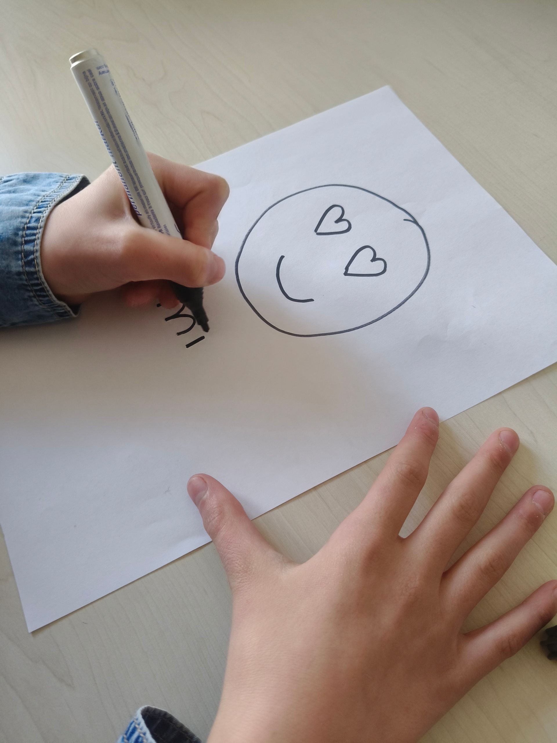 Een kind tekent met een stift een lachend gezichtje met hartjesogen en een bolle mond op wit papier.