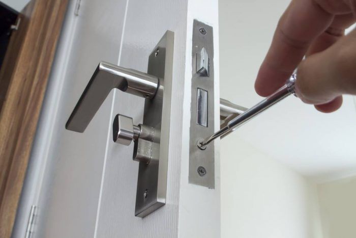A hand uses a screwdriver to tighten a screw on the metal faceplate of a door lock assembly on a white door.