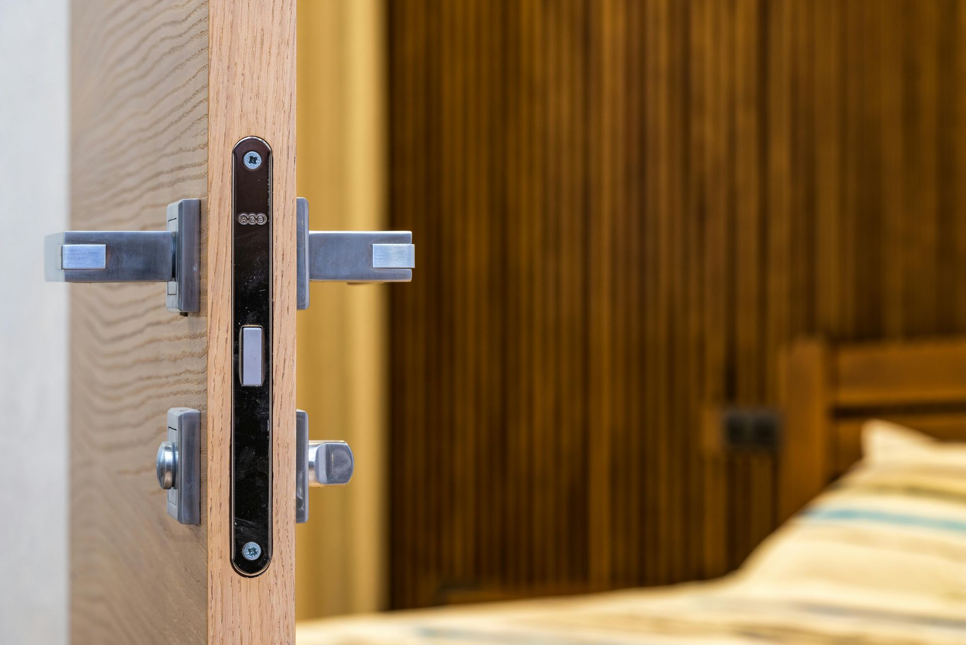 Close-up view of a light wood door edge featuring a metallic lock mechanism, with a blurred bedroom interior background.