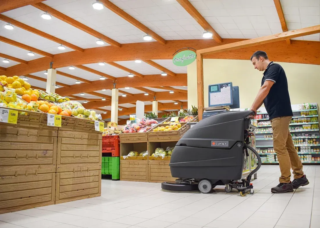 Person operating a floor cleaner in a grocery store, near produce displays.