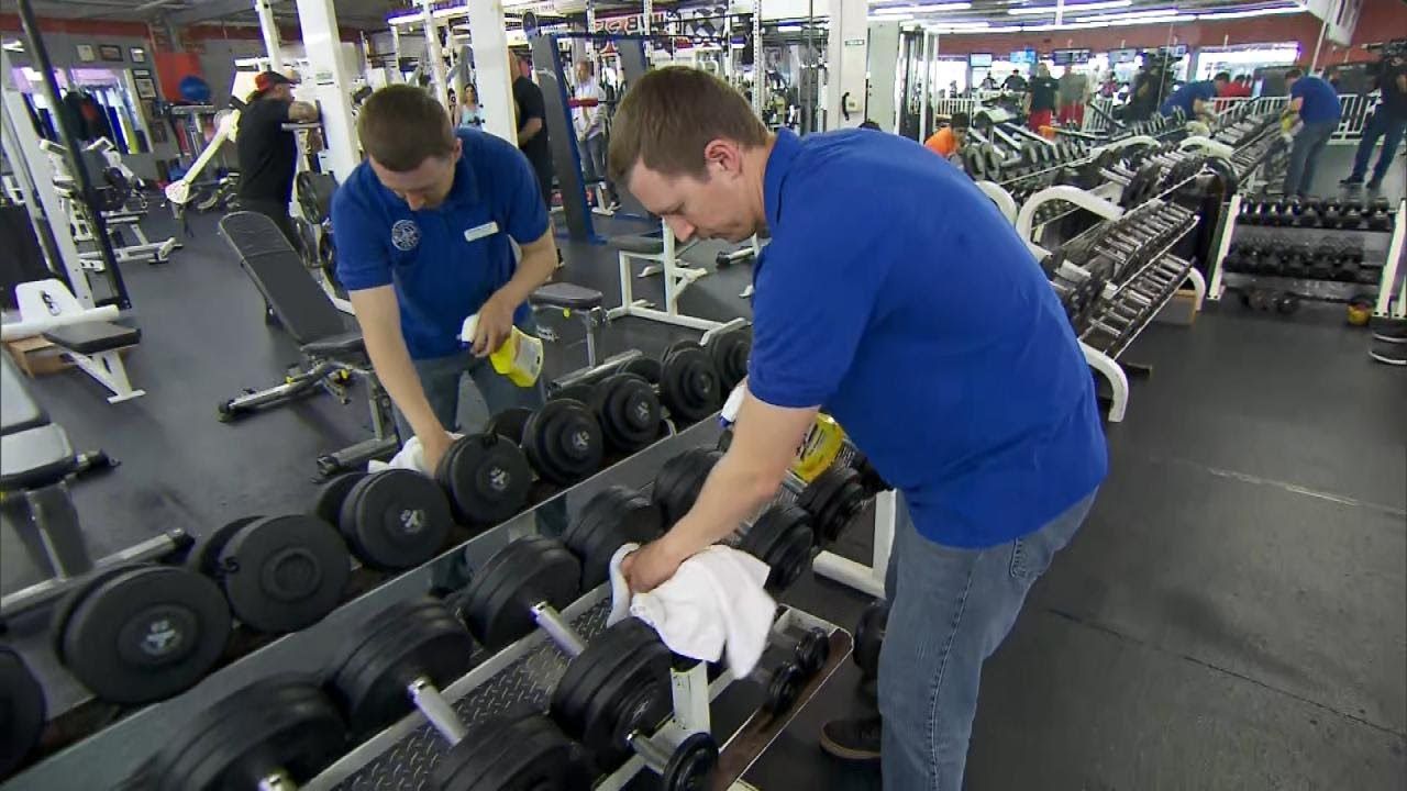 Two people in blue shirts cleaning weights with spray and cloth in a gym.