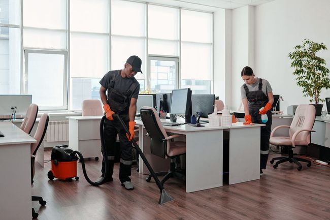 Two people cleaning an office: one vacuuming, the other wiping surfaces. Setting is a bright office with desks.