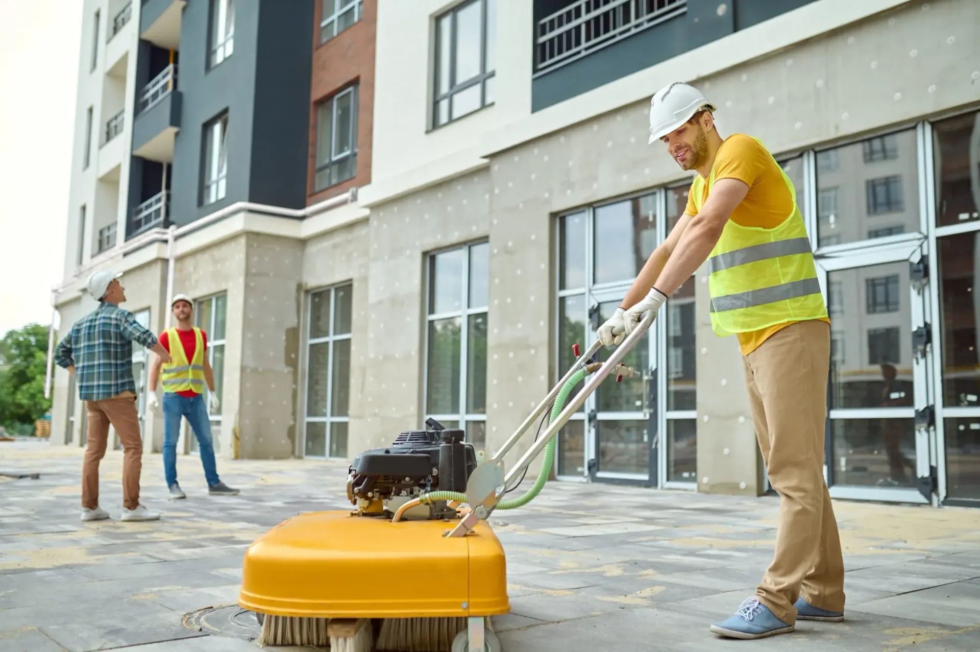 Construction worker operating a sidewalk sweeper, two others in background, next to building.