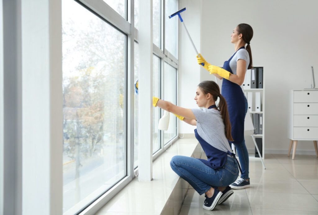 Two people cleaning large office windows with cleaning supplies.