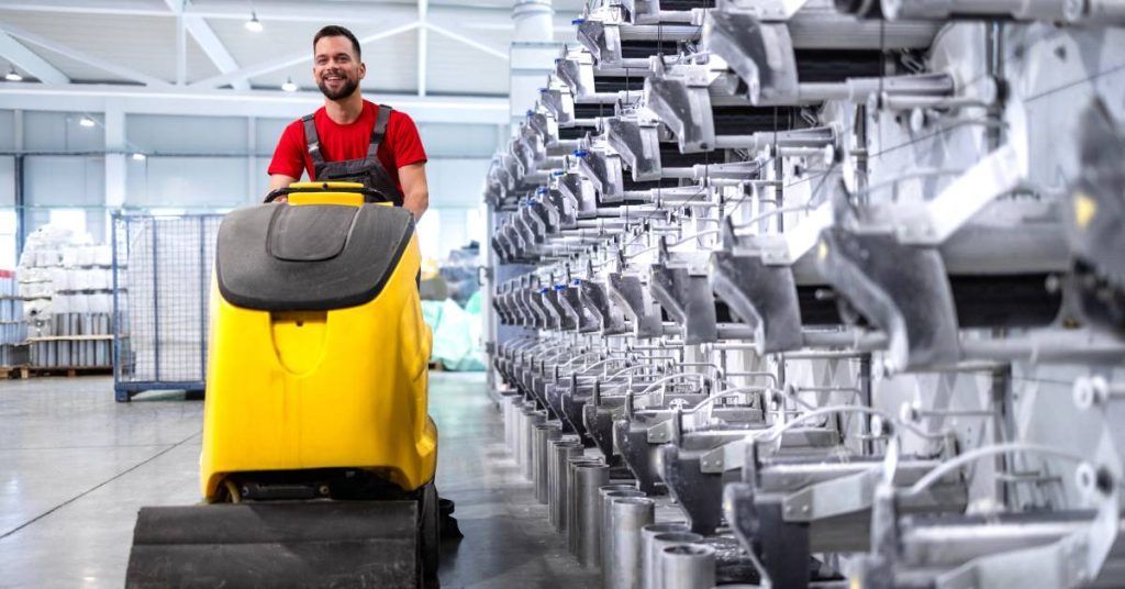 Man in red coveralls smiling, operating a yellow industrial vehicle in a factory setting.