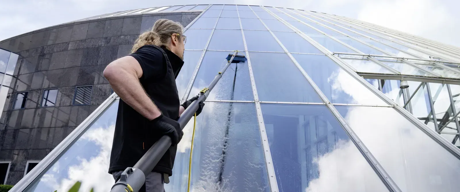 Person washes windows on a large glass building using a long-handled brush.