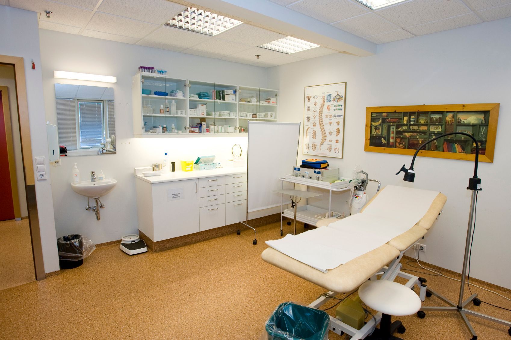 Doctor's examination room, light and airy. Examination table, cabinets, sink, medical equipment, and chart on the wall.