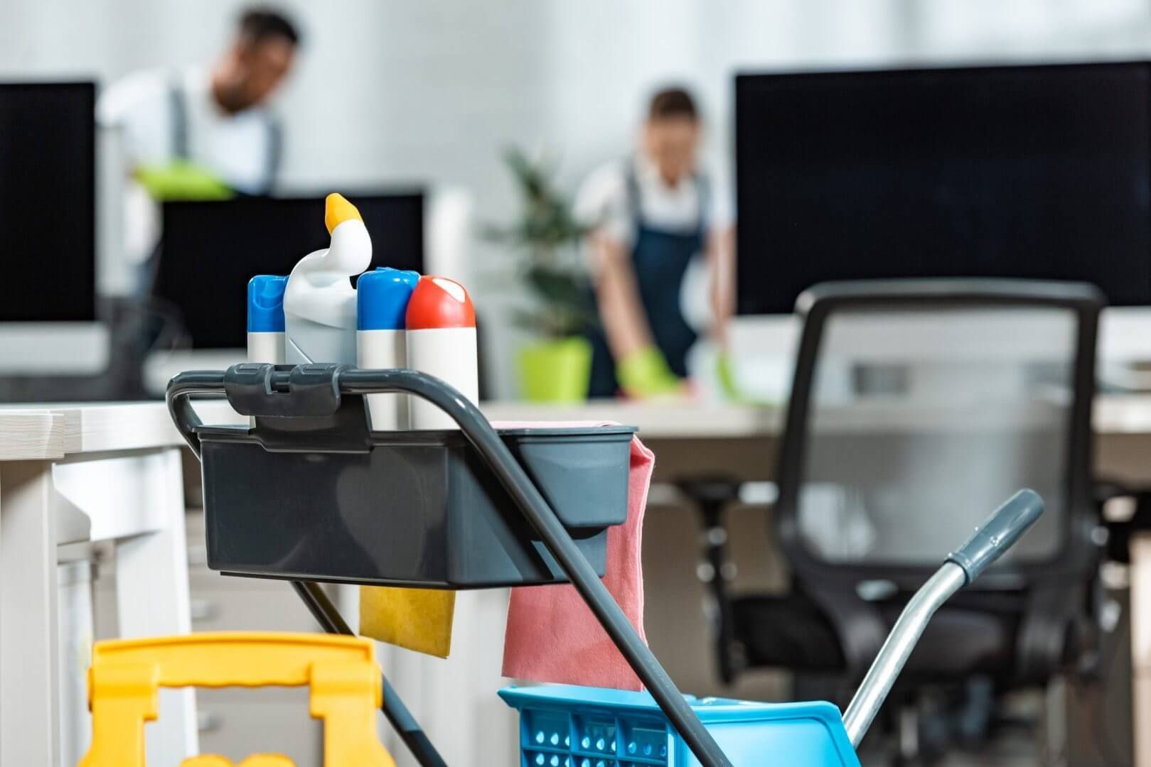 Cleaning cart in office; two workers cleaning desks in the background.