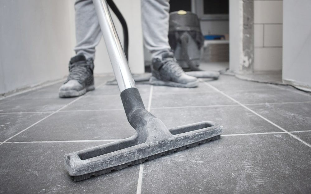 Person vacuuming dusty, grey tiled floor with a silver and black vacuum cleaner.