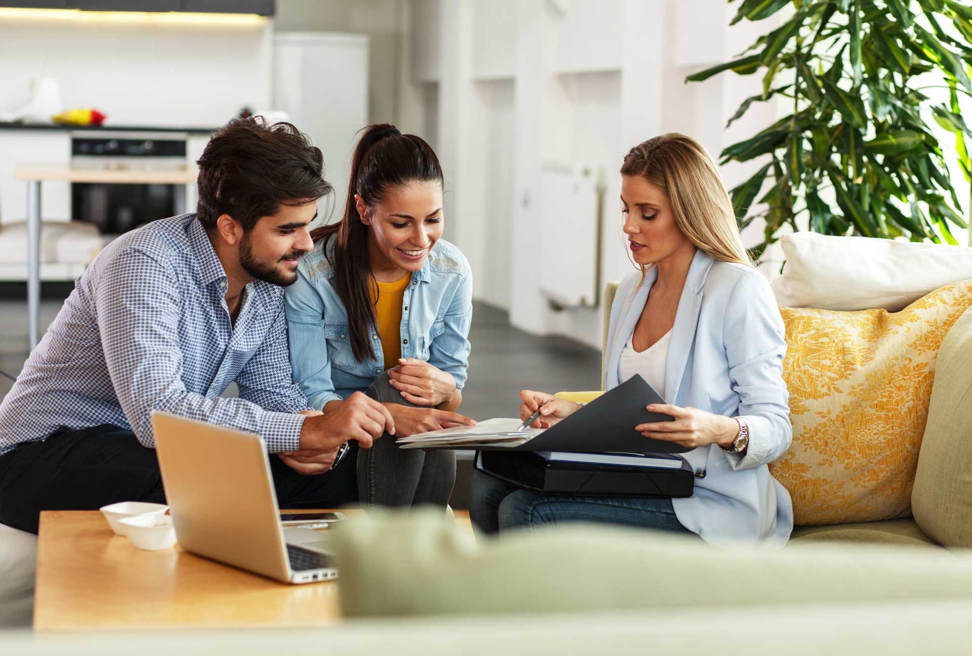 Couple and advisor reviewing documents in a living room, with a laptop and greenery nearby.