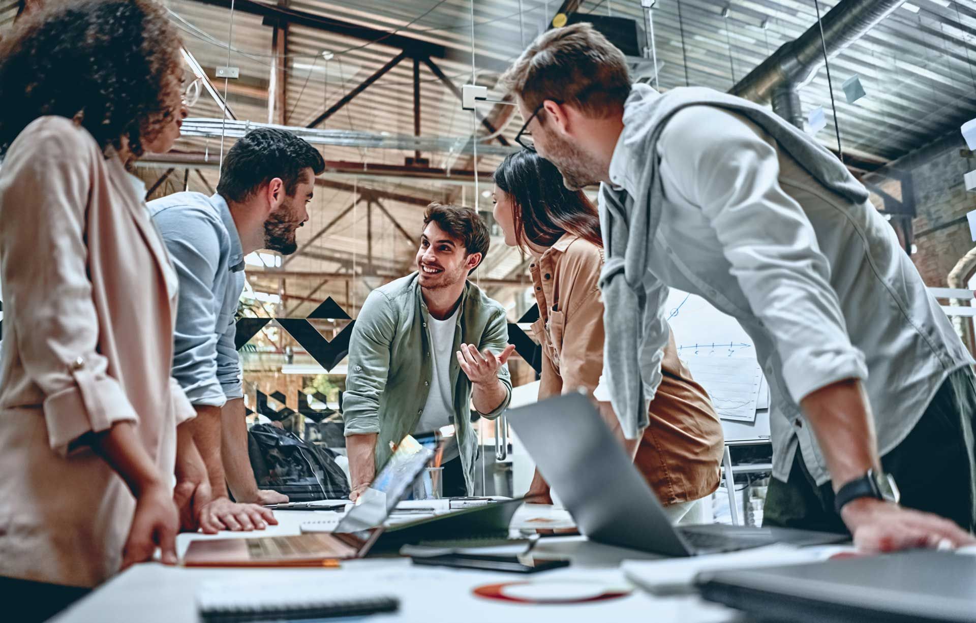 Group of people collaborating around a table with laptops, discussing ideas in a modern office space.