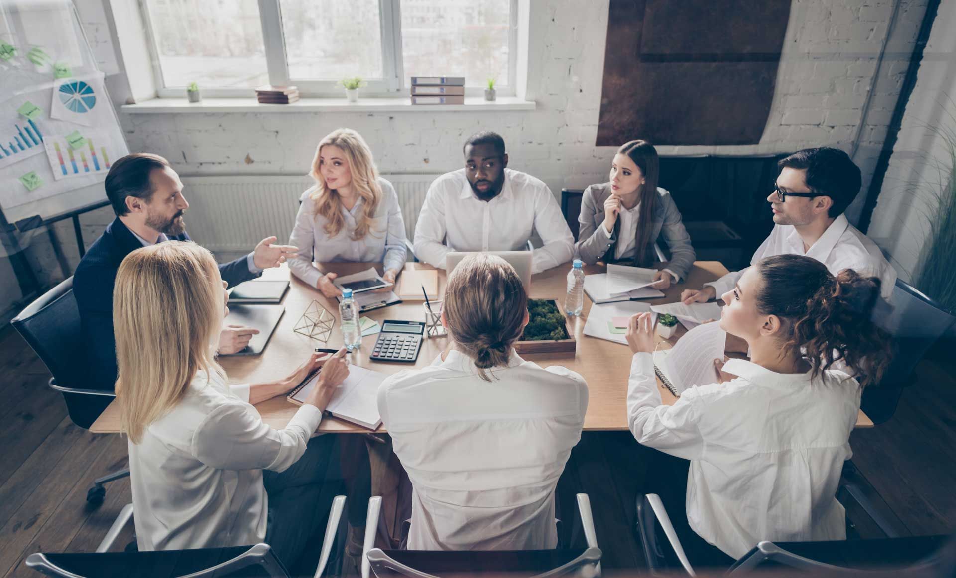 Group of professionals in a meeting, around a table with papers, whiteboard in background.
