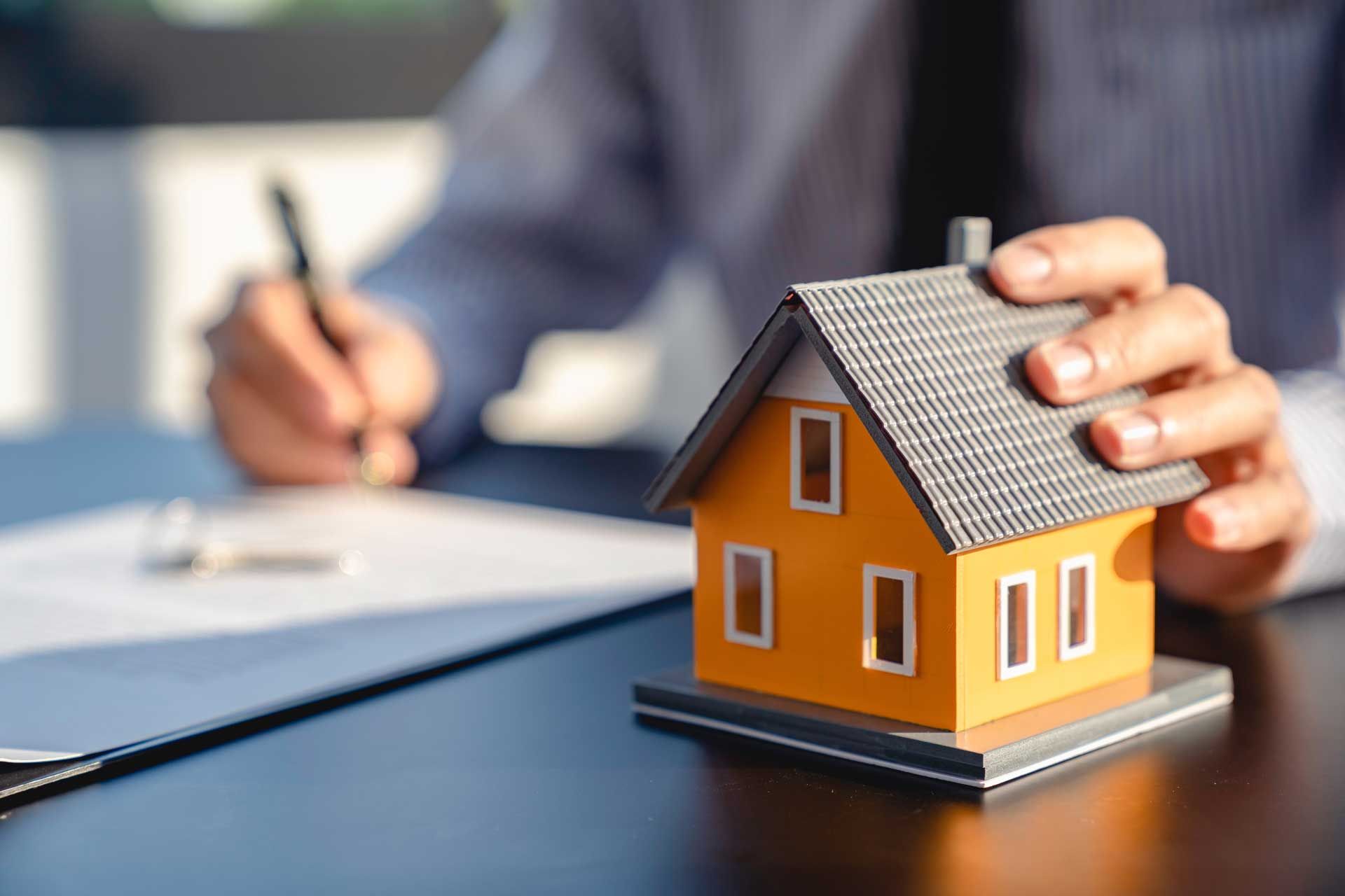 Person signing documents at a desk next to a miniature yellow house.