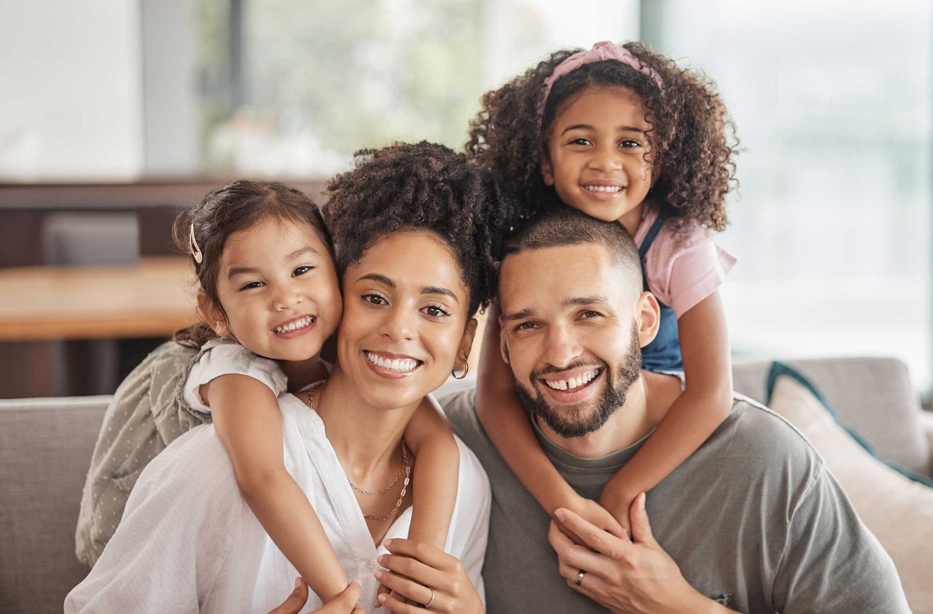Family of four smiling, posing indoors. Children on parents' shoulders, happy expressions.