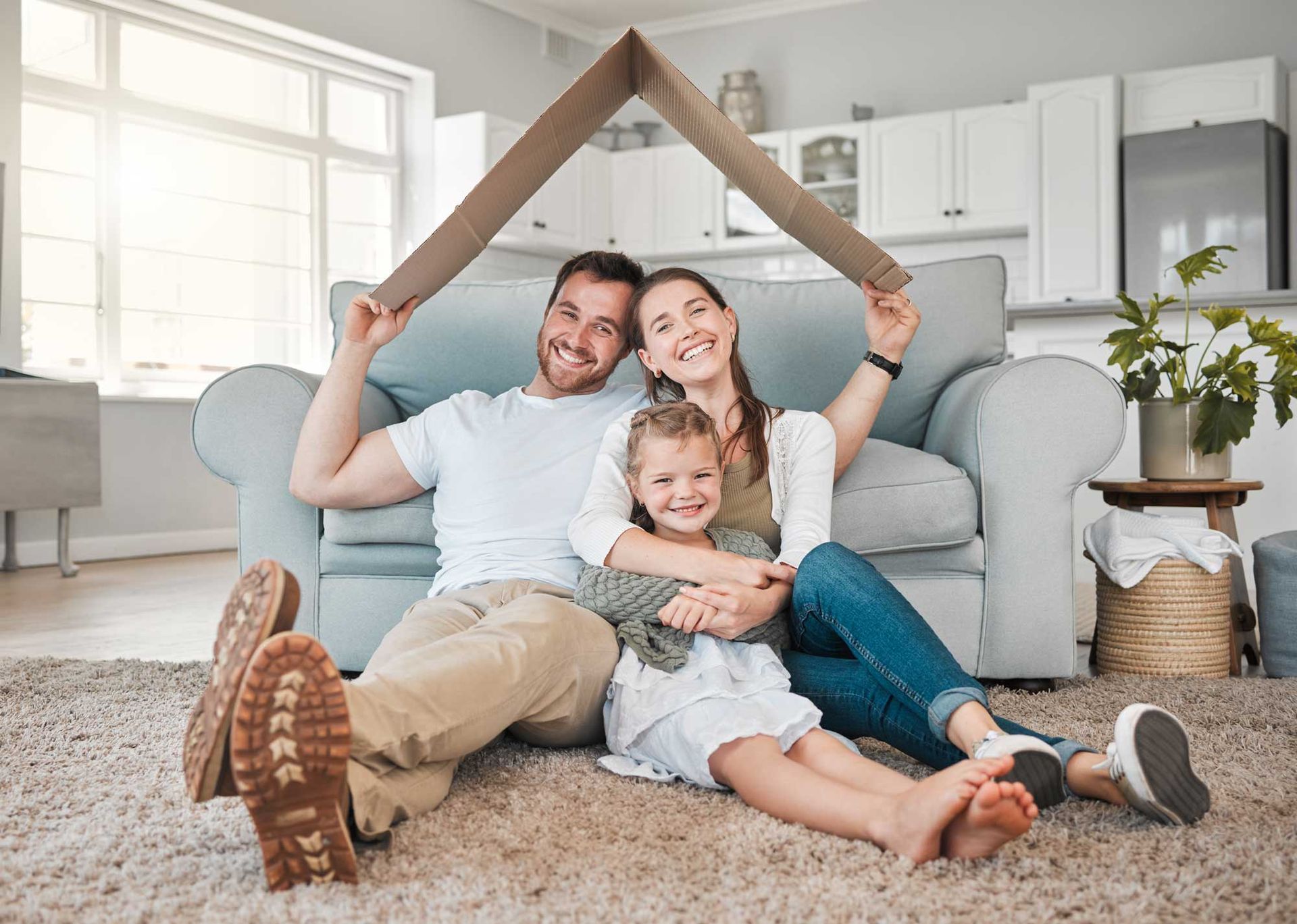 Family of three sits under a cardboard house indoors, smiling. Family of three sits under a cardboard house indoors, smiling.