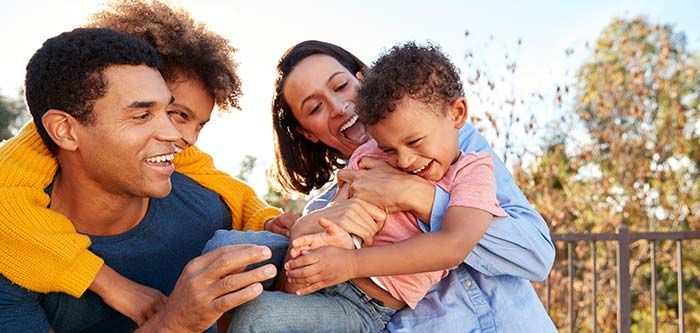 A family laughing and hugging outdoors; trees in the background.