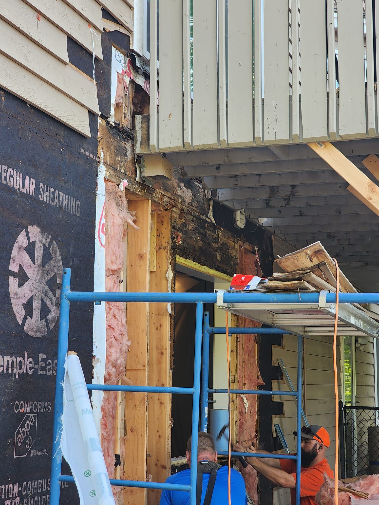 Construction site: Exterior wall partially open, revealing insulation, scaffolding, and two workers.