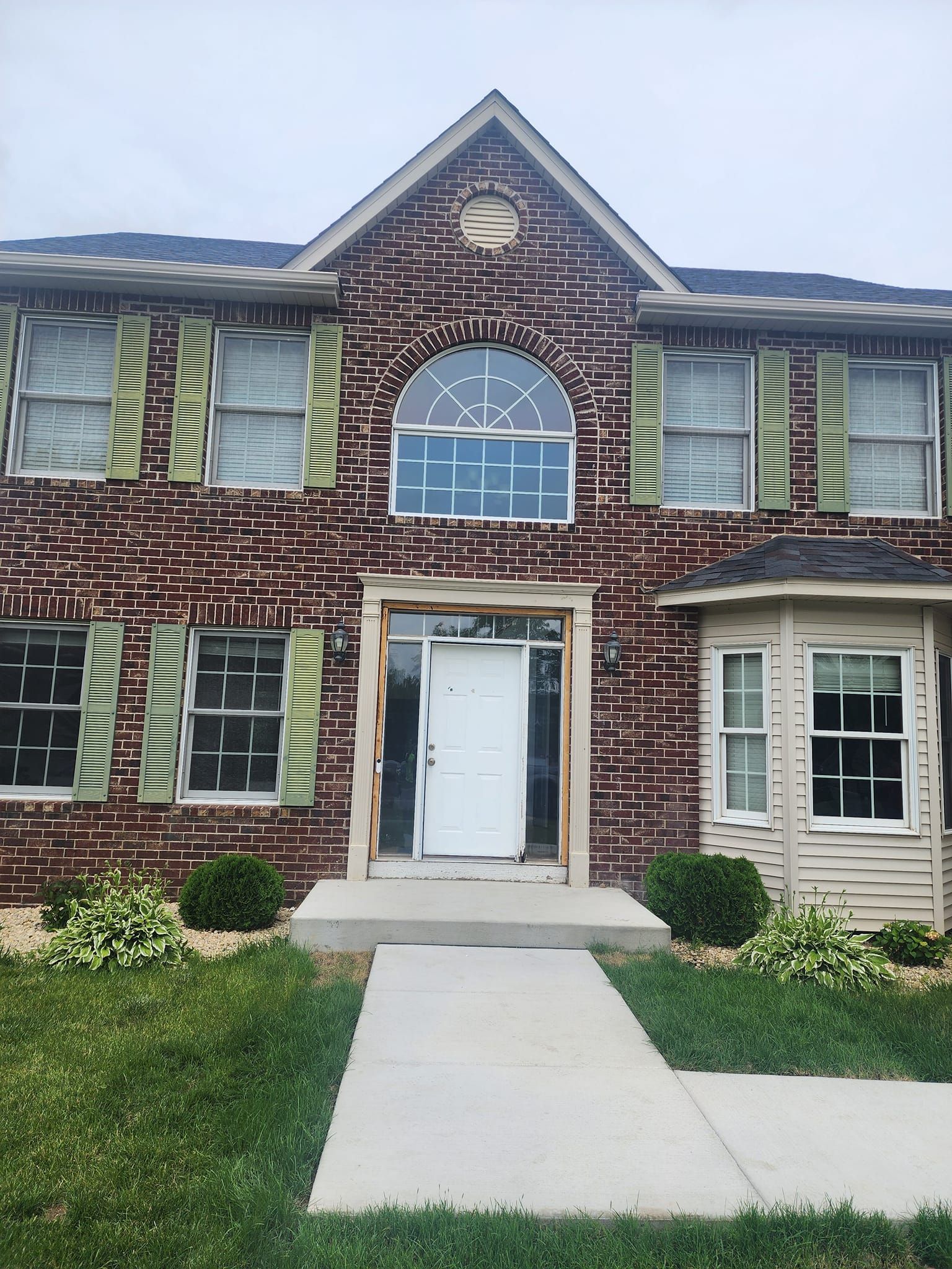 Two-story brick house with green shutters and a white door, centered on a concrete walkway.