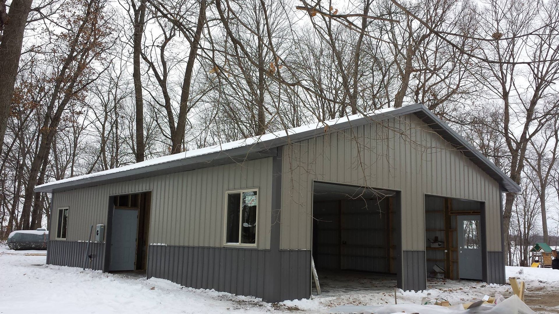 Metal garage building with gray and silver siding, snowy ground, and bare trees.