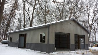 Gray and silver metal-sided garage with two bays, two doors, and windows, set in a snowy landscape with bare trees.