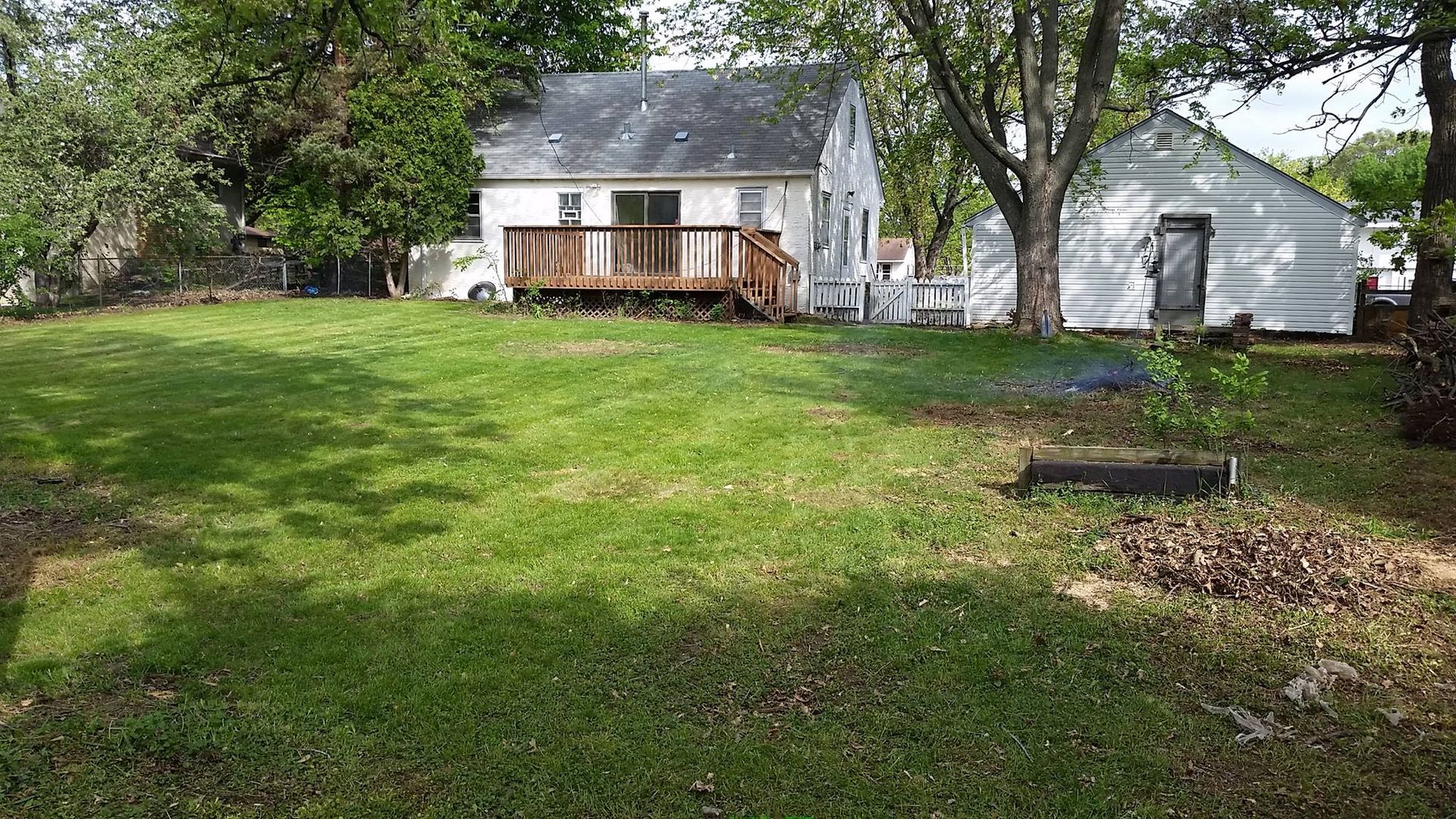 A grassy backyard with a house, deck, and detached garage; trees surround the perimeter.