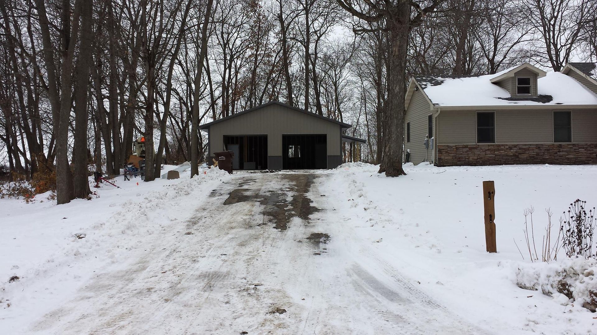 Snowy driveway leading to a garage with two doors. A house is on the right.