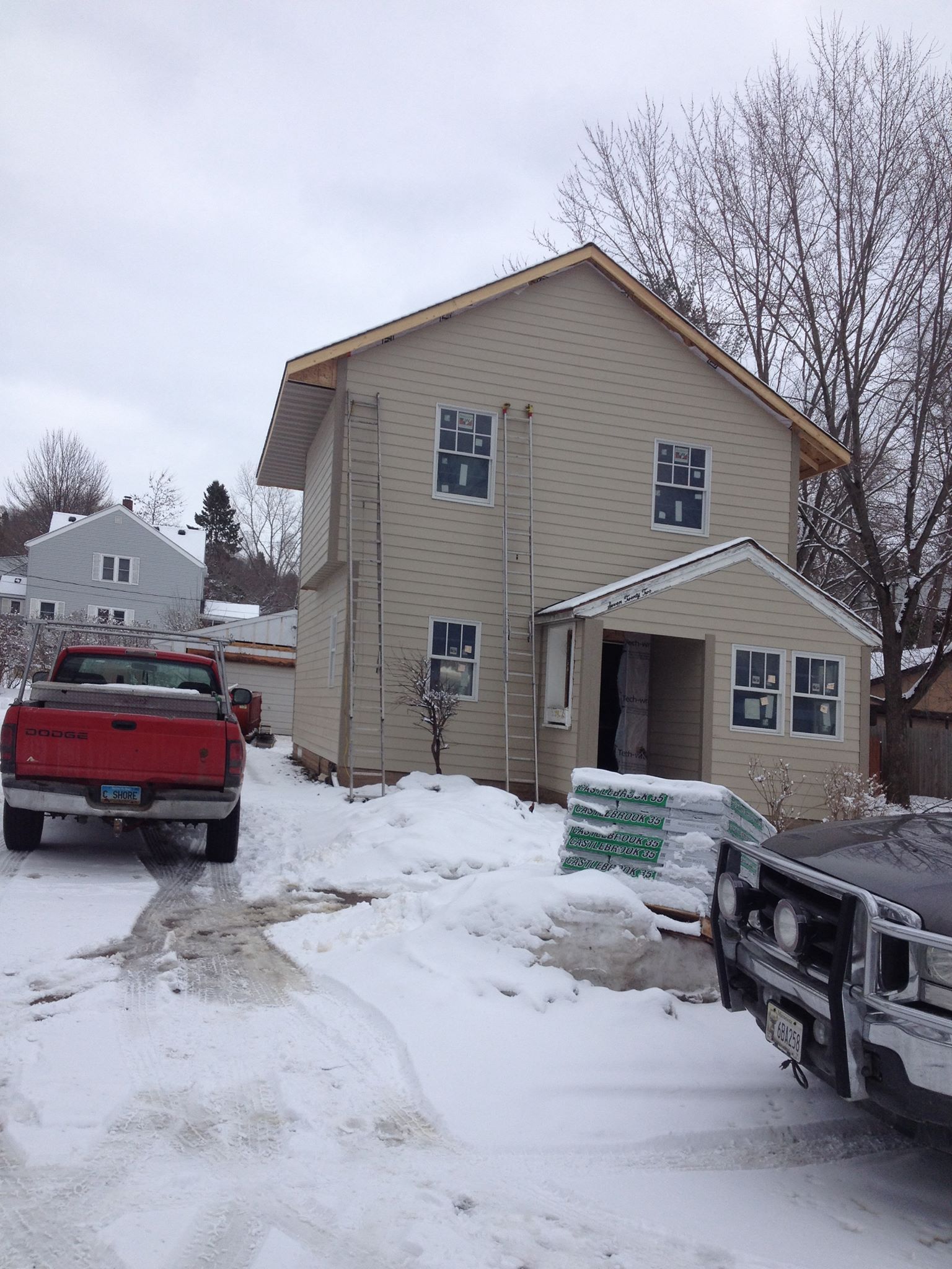 Two-story house with tan siding, under construction, with snow on the ground and parked vehicles.