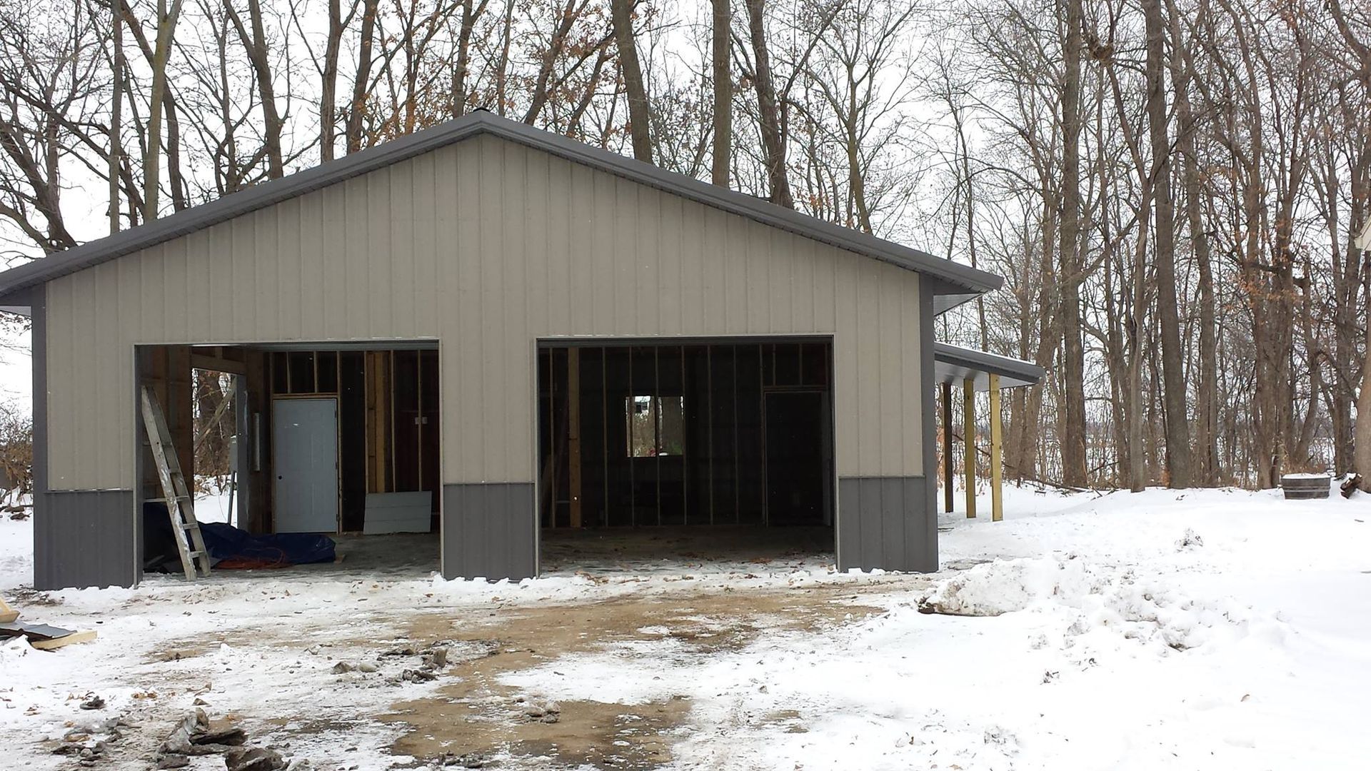 Metal-sided garage under construction in a snowy setting with two open bays and a gray roof.