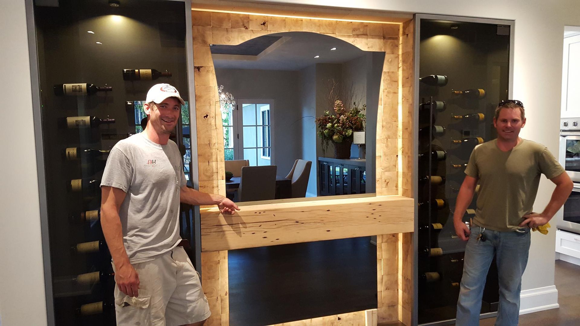 Two men standing by a wine rack with a mirror. Wooden frame with lights, a dark interior, and a light-filled background.
