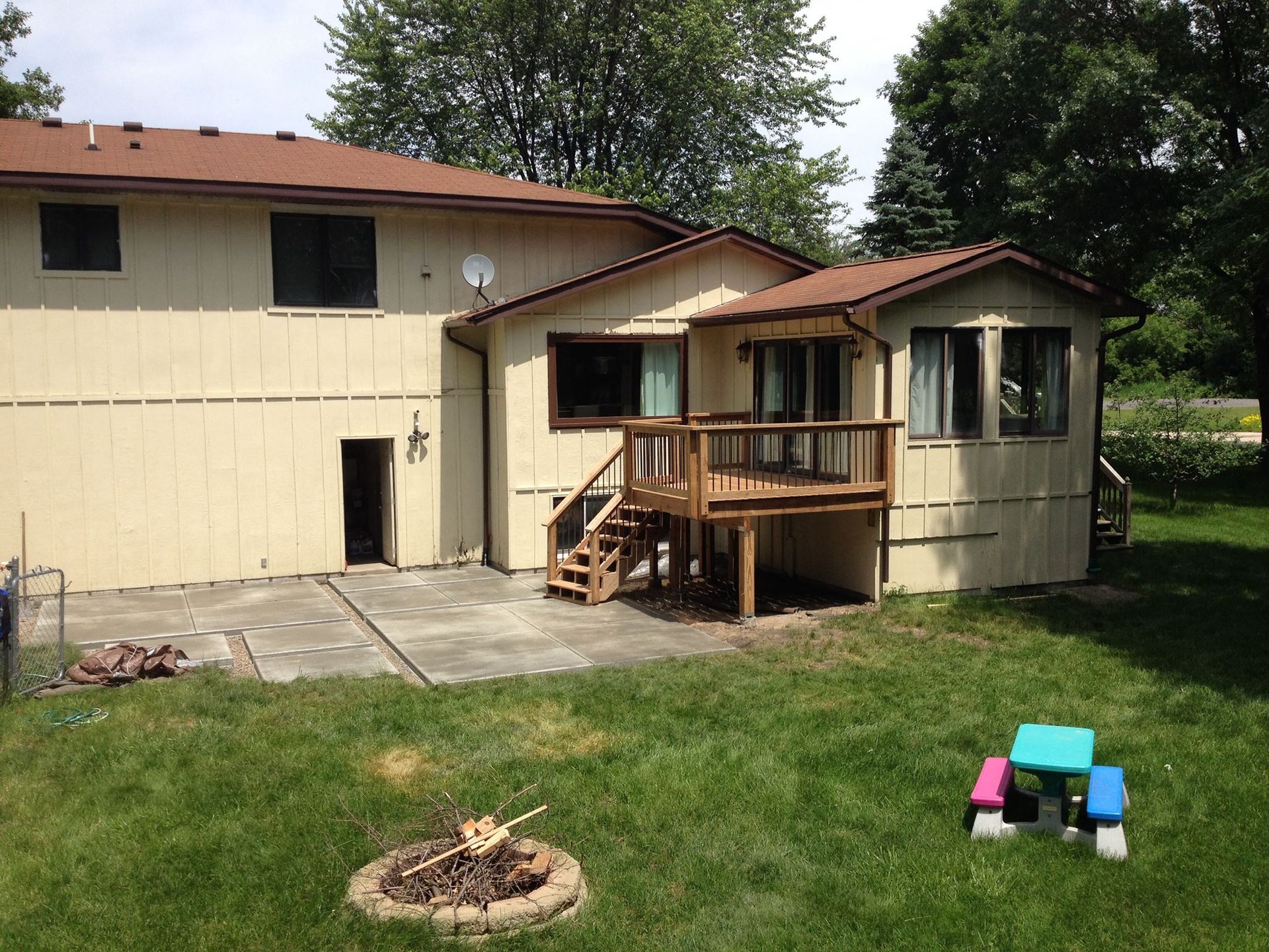 Backyard with a light yellow house, brown roof, and wooden deck. A fire pit and small table sit on the grass.