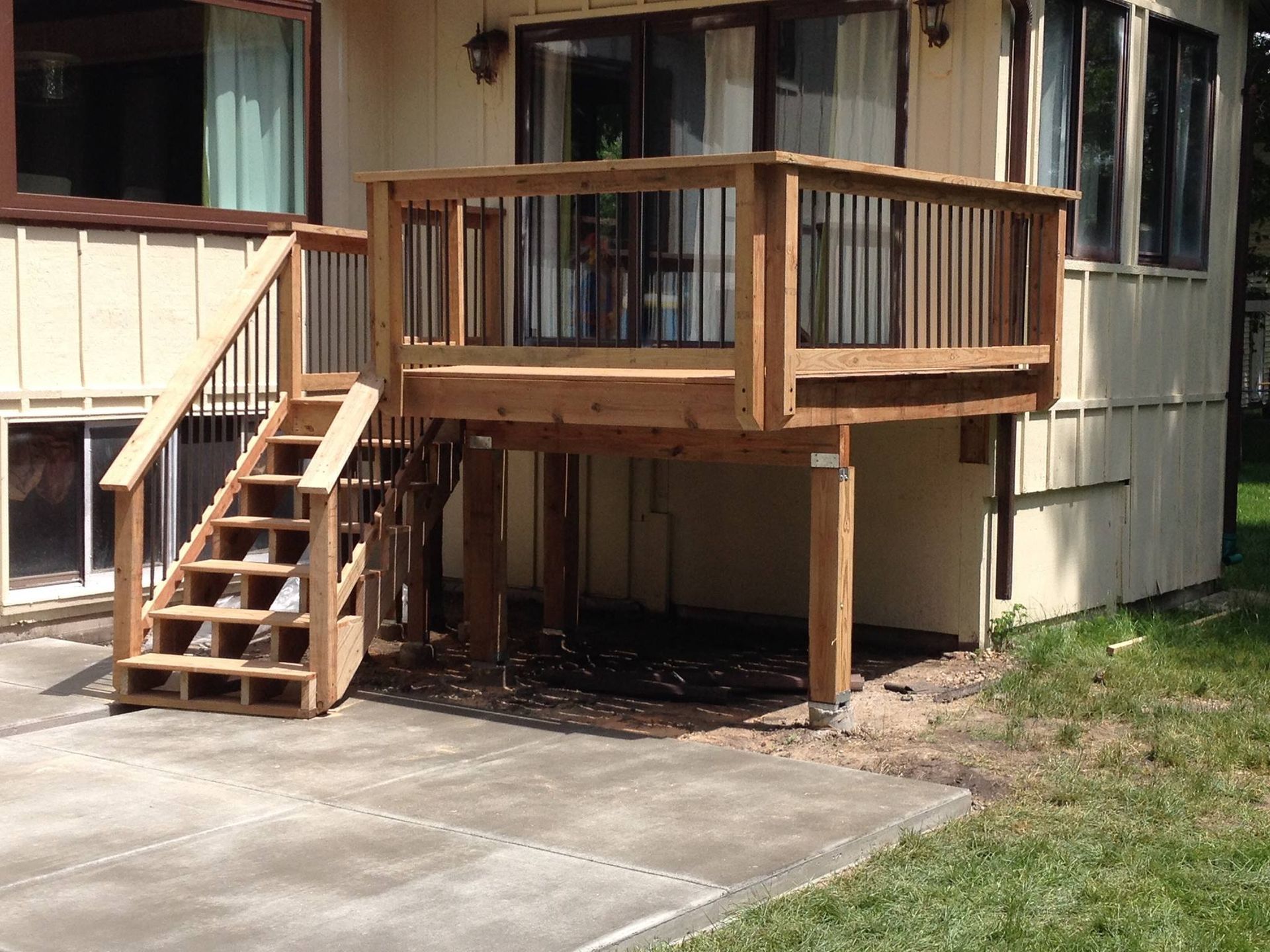 Wooden deck with stairs, built against a house with a concrete patio and grass.