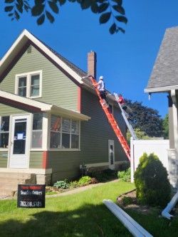 Man on a ladder repairing a roof on a green house. A lawn is in the front with sunny sky.