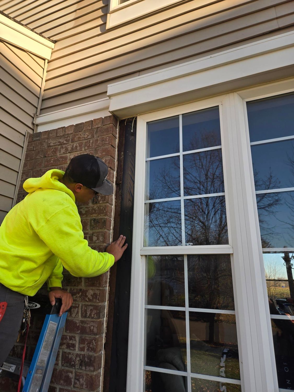 A person in a yellow jacket works on a window frame, exterior brick wall.