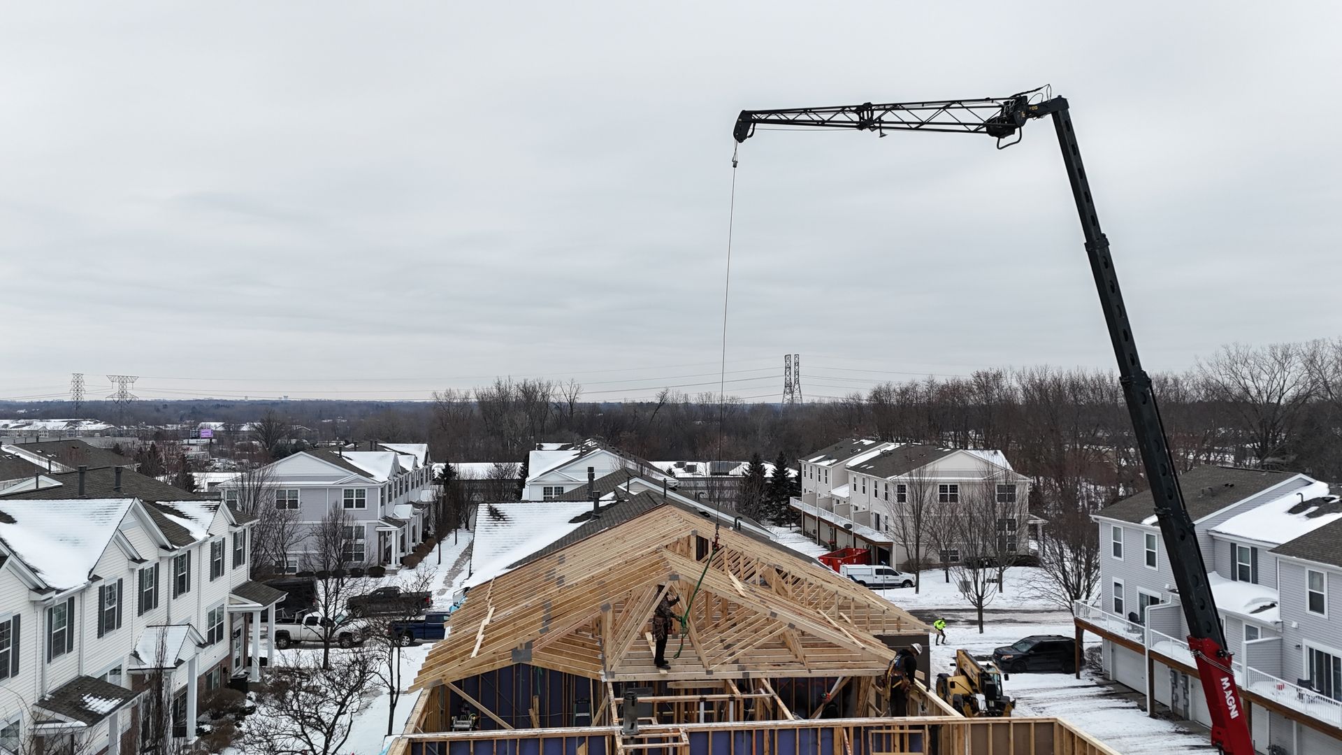 Construction of a house with a crane lifting materials on a snowy day. Houses and trees in the background.