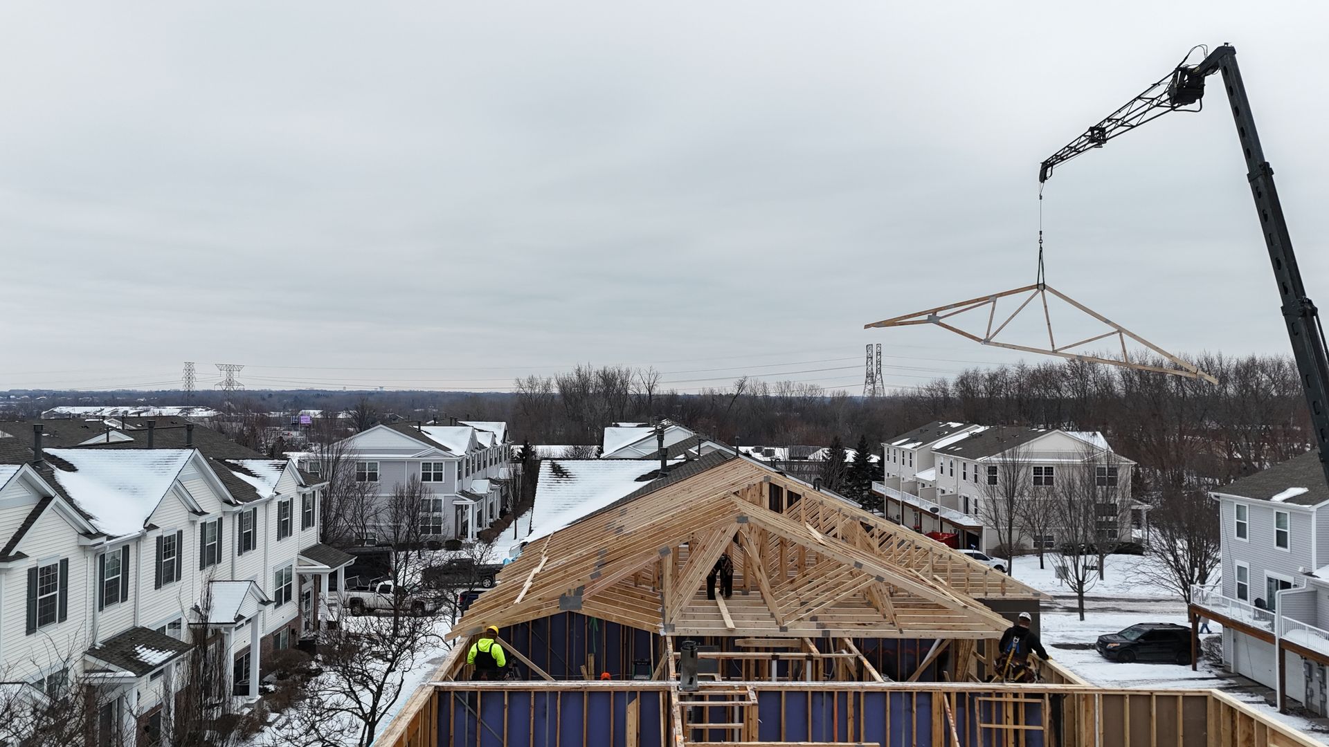 Construction site, crane lifting a roof truss over a partially built house, winter setting, workers present.