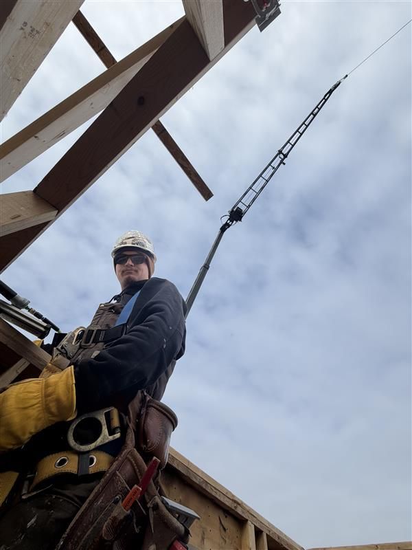 Construction worker on roof with safety harness, next to a tall pole, cloudy sky.