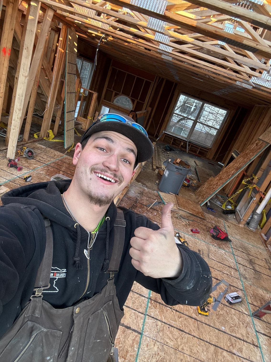 Man in work clothes gives thumbs up inside a construction site. Wooden beams and tools are visible.