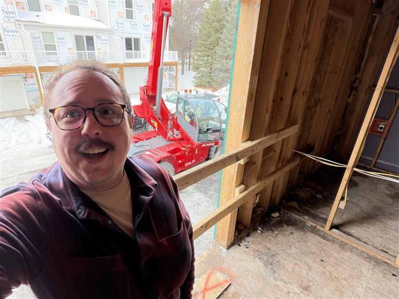 Man in glasses smiles at camera near construction site with a red forklift and building frame.