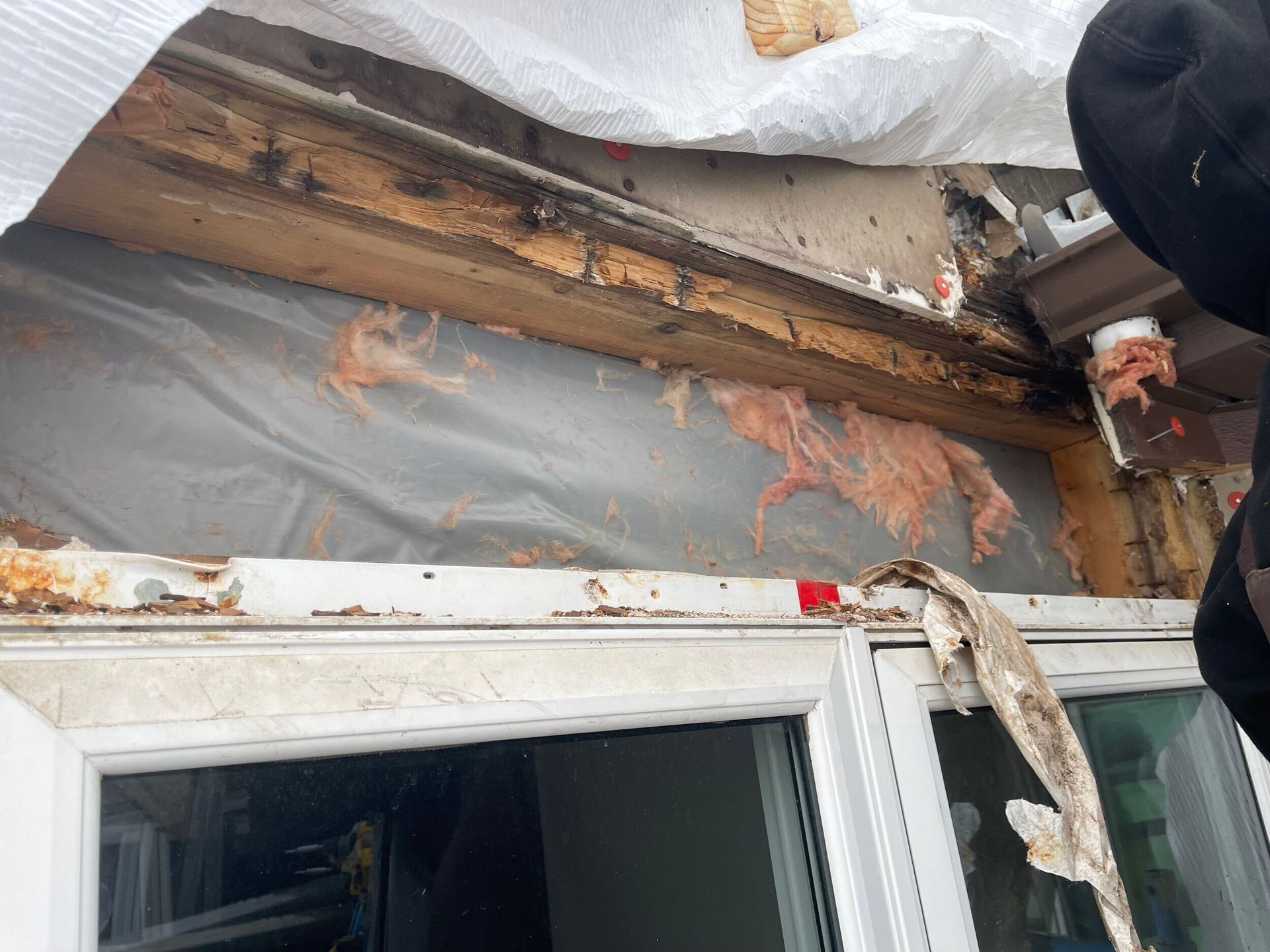 Damaged window frame with water damage and insulation exposed, covered by plastic sheeting.