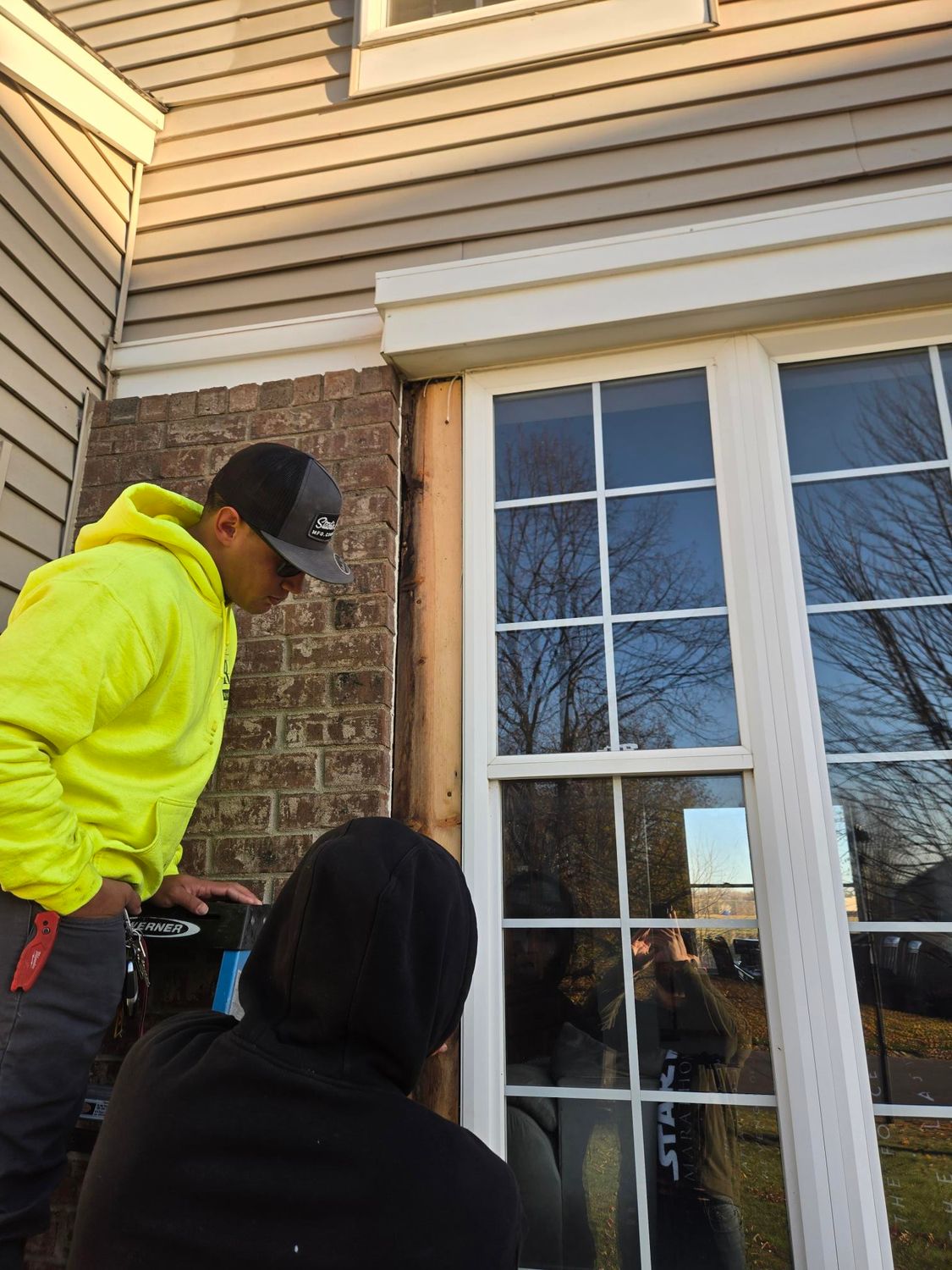 Two workers installing trim around a window on a brick and siding house. One wears a yellow hoodie.
