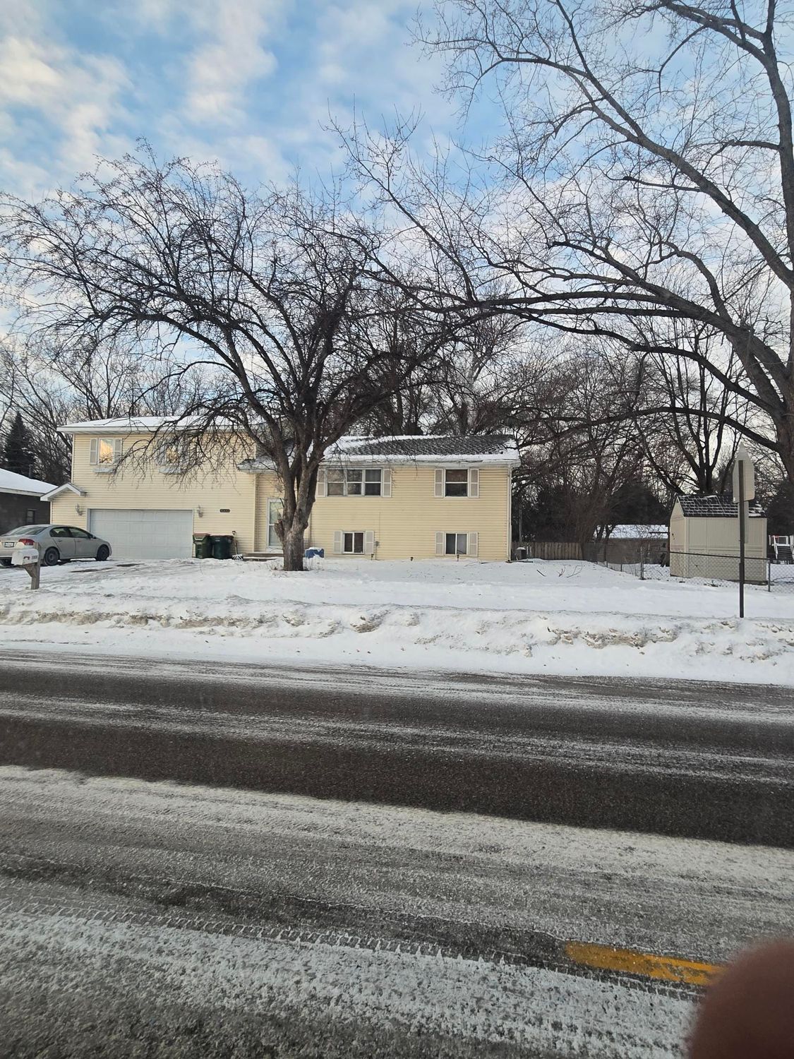 Yellow house with snow-covered yard and bare trees. A car is parked in the driveway; a road is in the foreground.