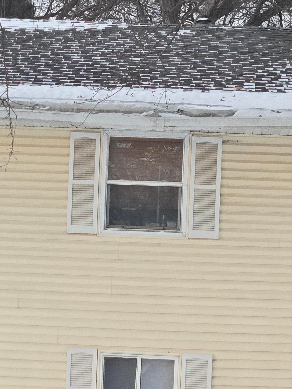 Yellow house with weathered white shutters and window under a snow-covered roof.