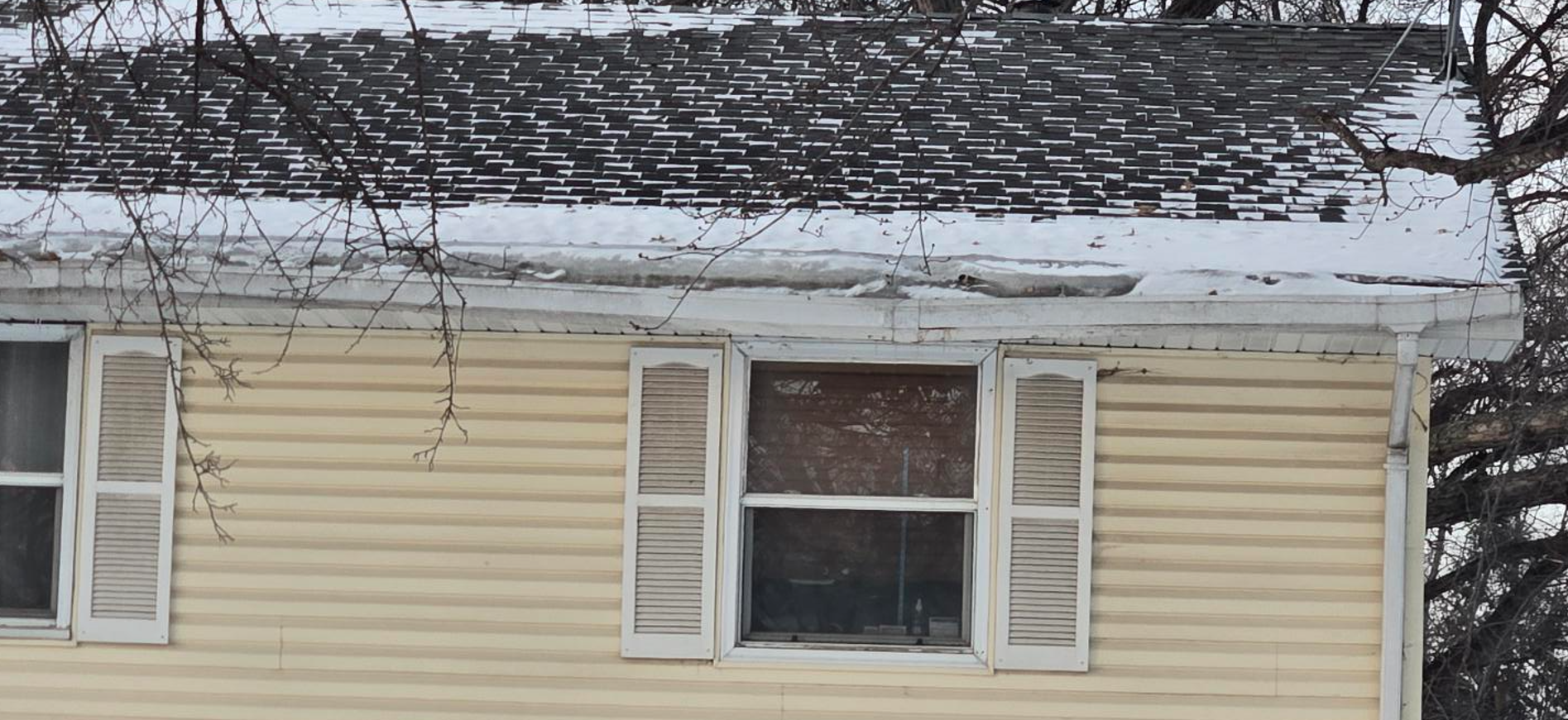 Snow-covered roof of a light yellow house with white shutters. Leafless tree branches overhead.