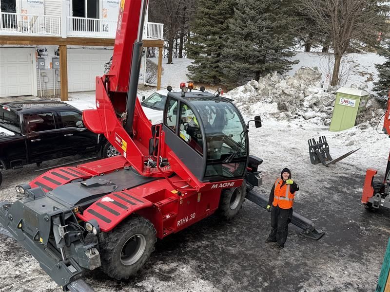 A red telehandler machine next to a person in an orange vest, winter scene.