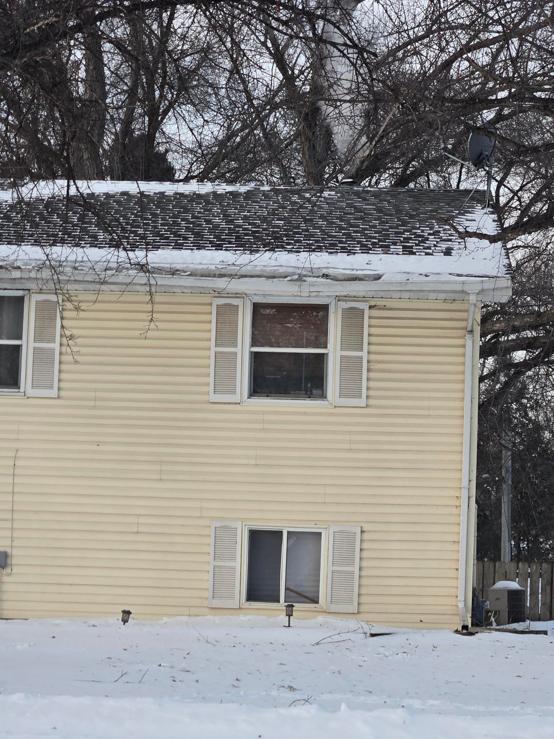 Yellow house with white shutters, snow on the ground and roof, bare trees in the background.